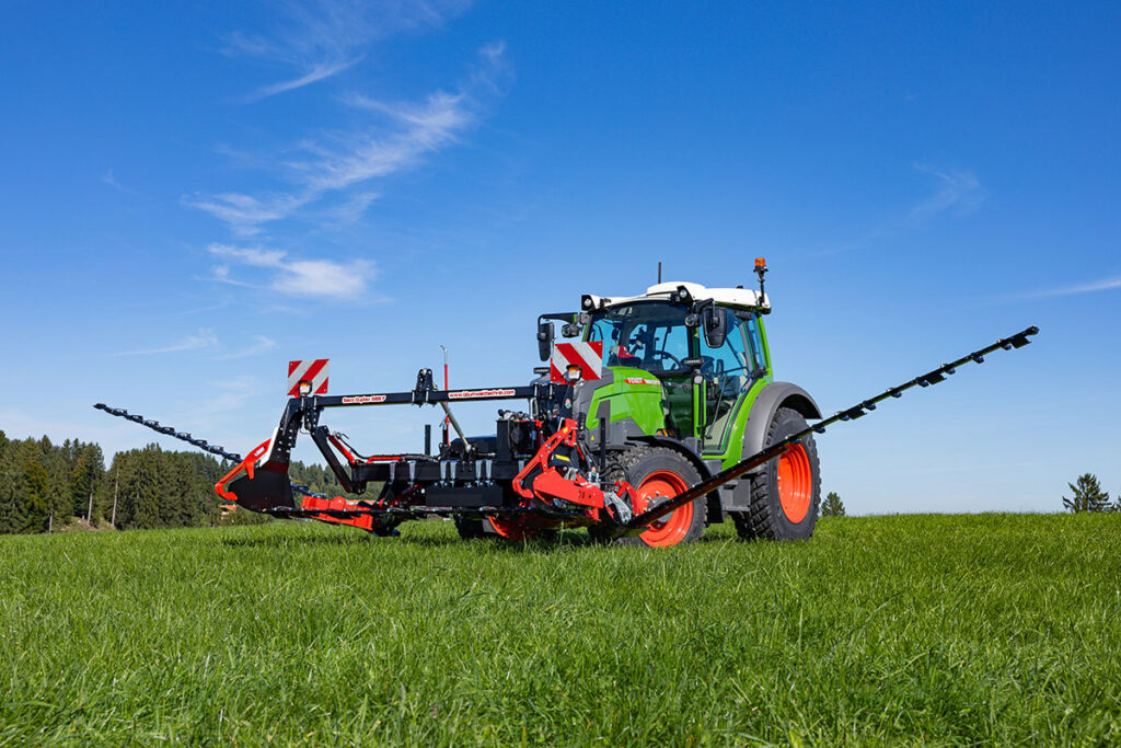 Frontschmetterlingsmaehwerk von BB-Umwelttechnik vor einem Fend Traktor auf gruener Wiese.